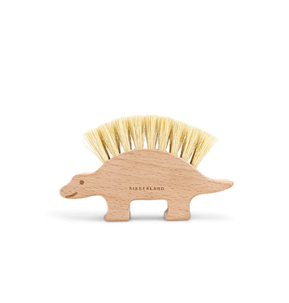 Wooden dinosaur-shaped brush with bristles extending from its back has a smiley face design and is stamped with the brand name, "Kikkerland"