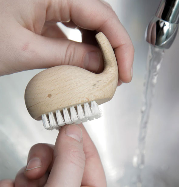 Model uses a wooden whale nail brush over a bathroom sink with running water