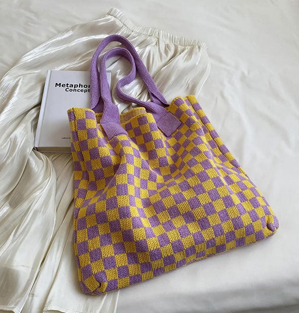 Knit yellow and purple checkered tote bag with purple straps rests on a white bedspread with a white satin skirt and Metaphorical Concepts book