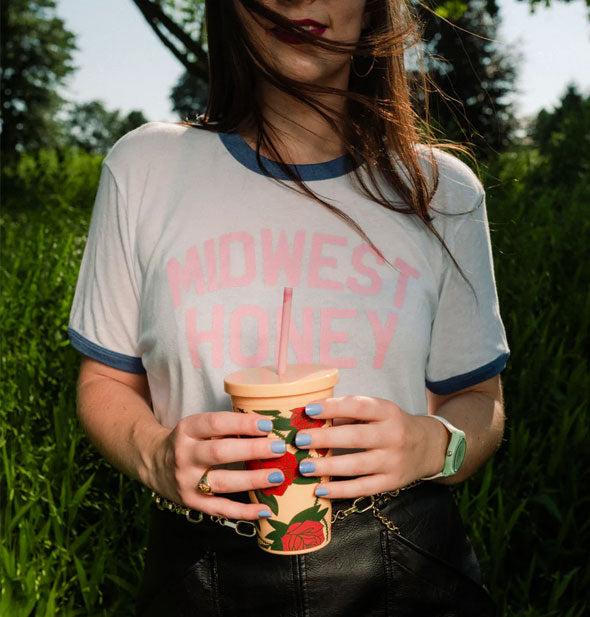 Model wearing "Midwest Honey" ringer T-shirt holds a yellow thorny rose drink tumbler against a background of field and trees