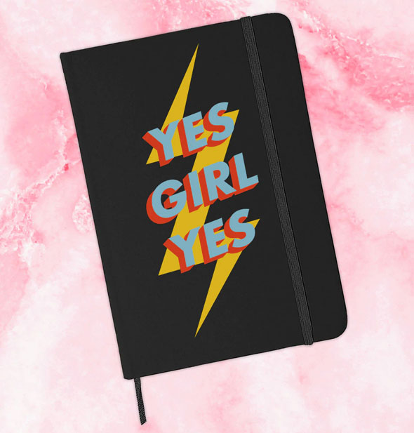Black journal with elastic band and ribbon place marker on a pink marbled backdrop says, "Yes Girl Yes" with lightning bolt graphic in primary colors