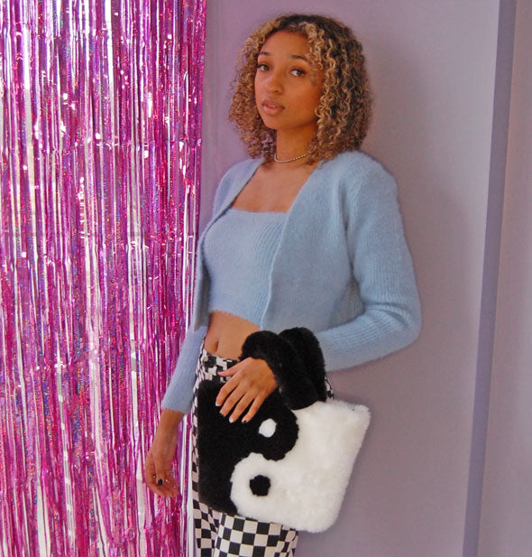 Model standing next to pink streamers holds a black and white fuzzy yin yang purse over left wrist