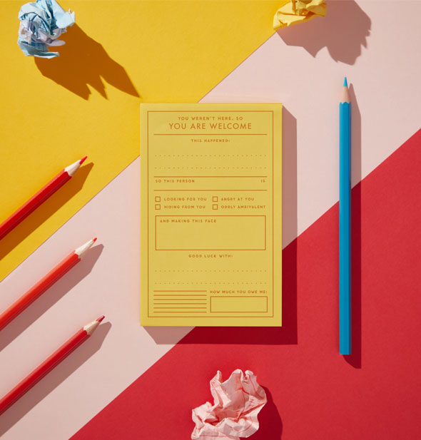 Yellow "You Are Welcome" notepad on a color blocked surface with blue and red pencils and crumpled-up pieces of paper