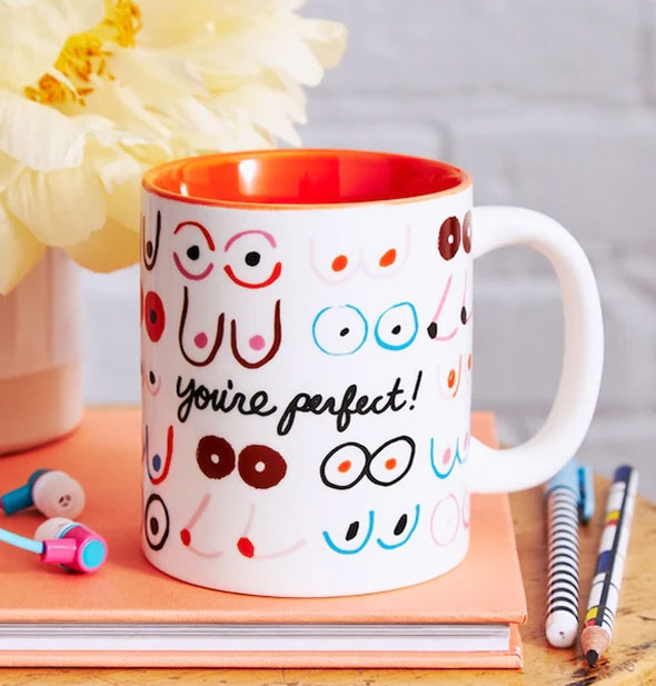 You're Perfect! Boobies mug rests on a book with flowers, earbuds, and pencils
