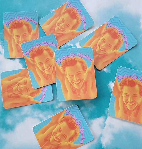 Grouping of rectangular stickers that feature a monochromatic orange image of Harry Styles below the words, "You're So Golden" in pink script, arranged on a background of blue sky with white clouds